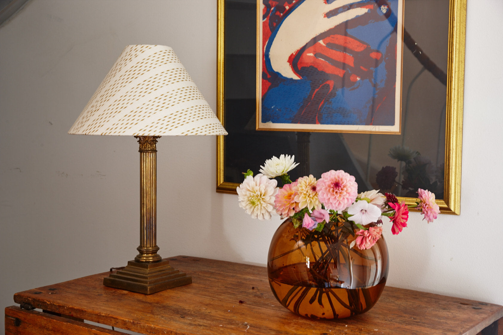 A table lamp with a shade in cream and gold on a brass column, on a wooden table beside a vase of dahlias