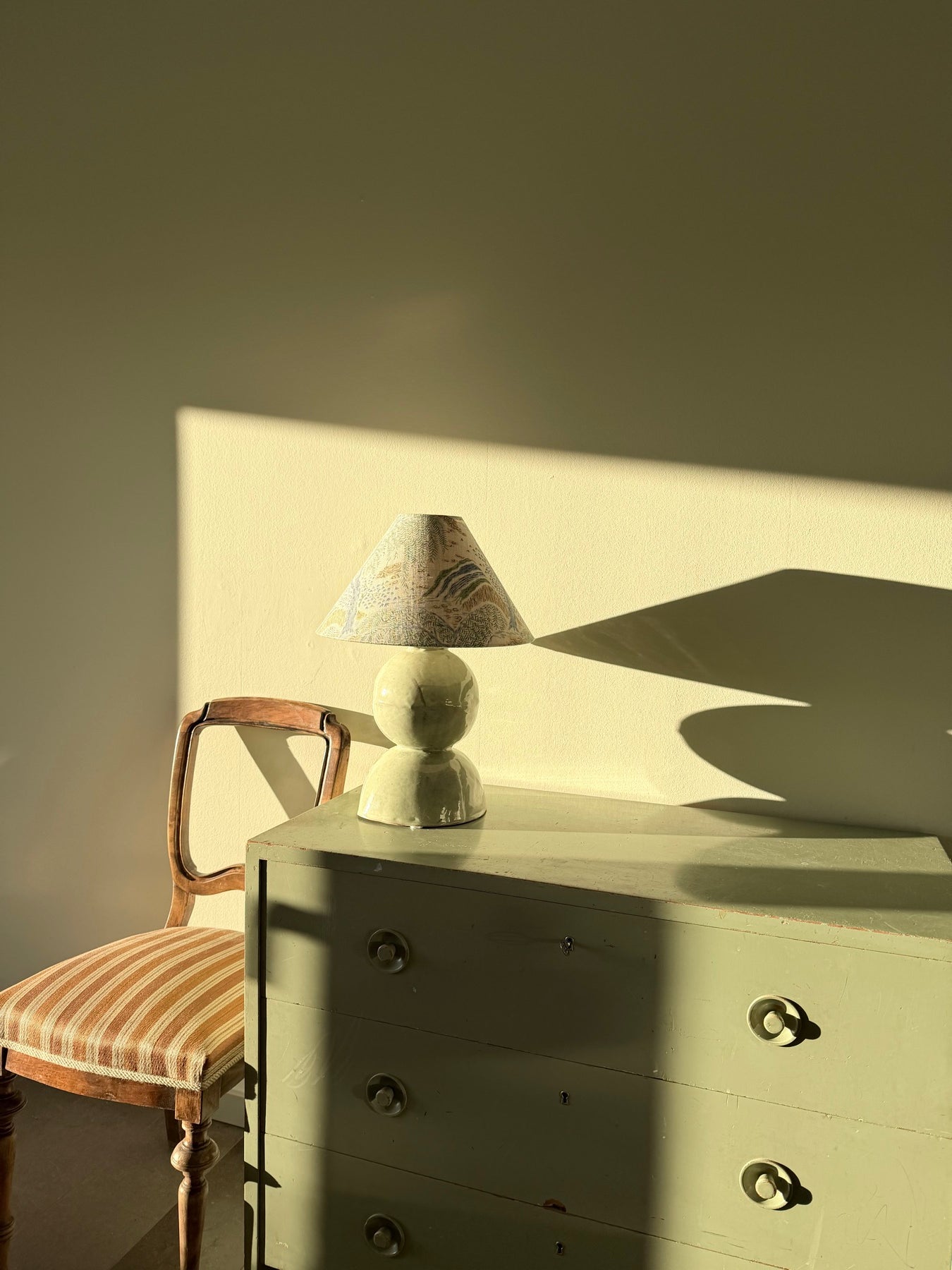 A lampshade on a green painted chest of drawers, lit by afternoon sun