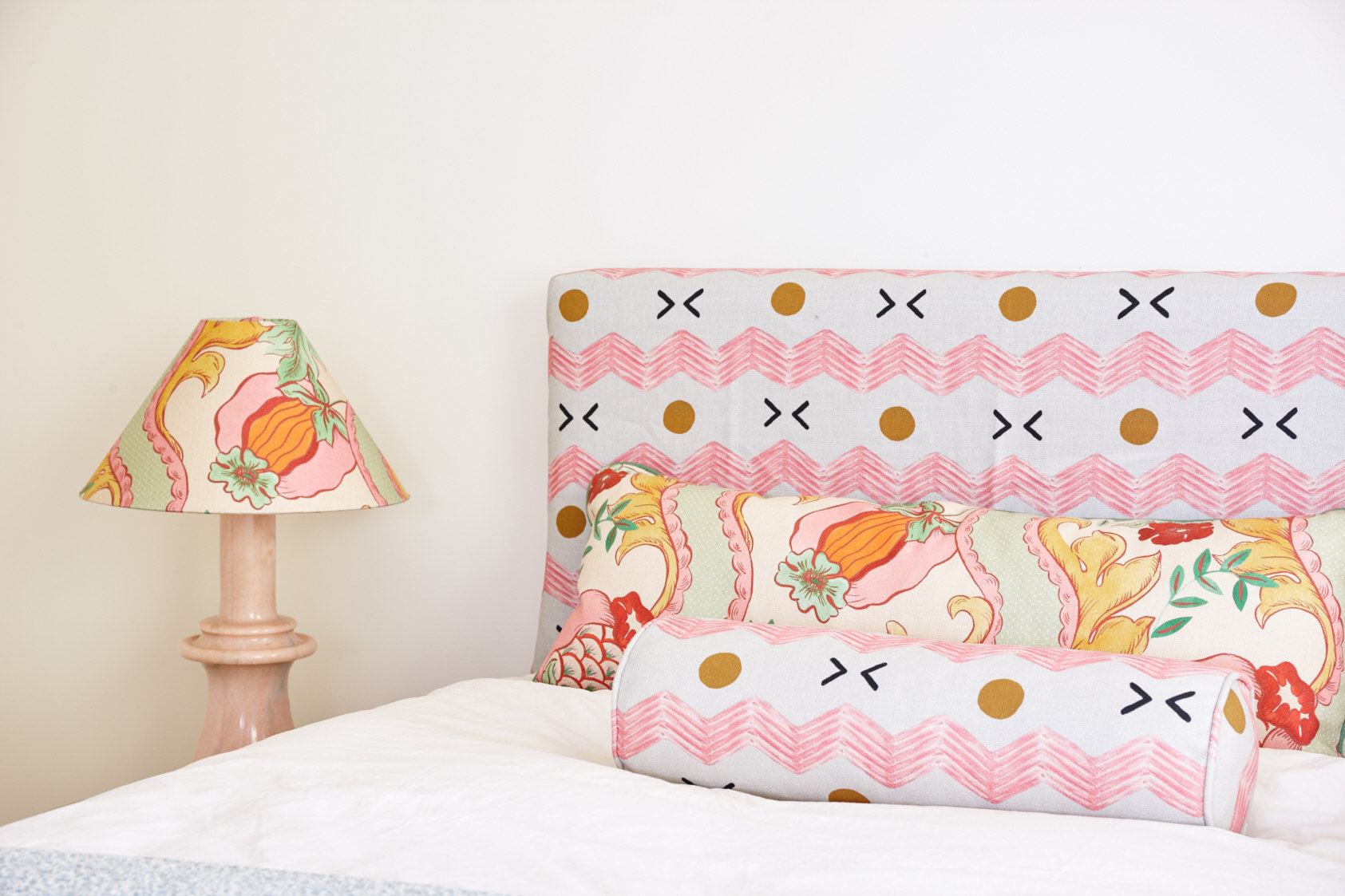 A bolster and a square cushion in printed cottons on a bed, against an upholstered headboard, with a lampshade in the same fabric beside it