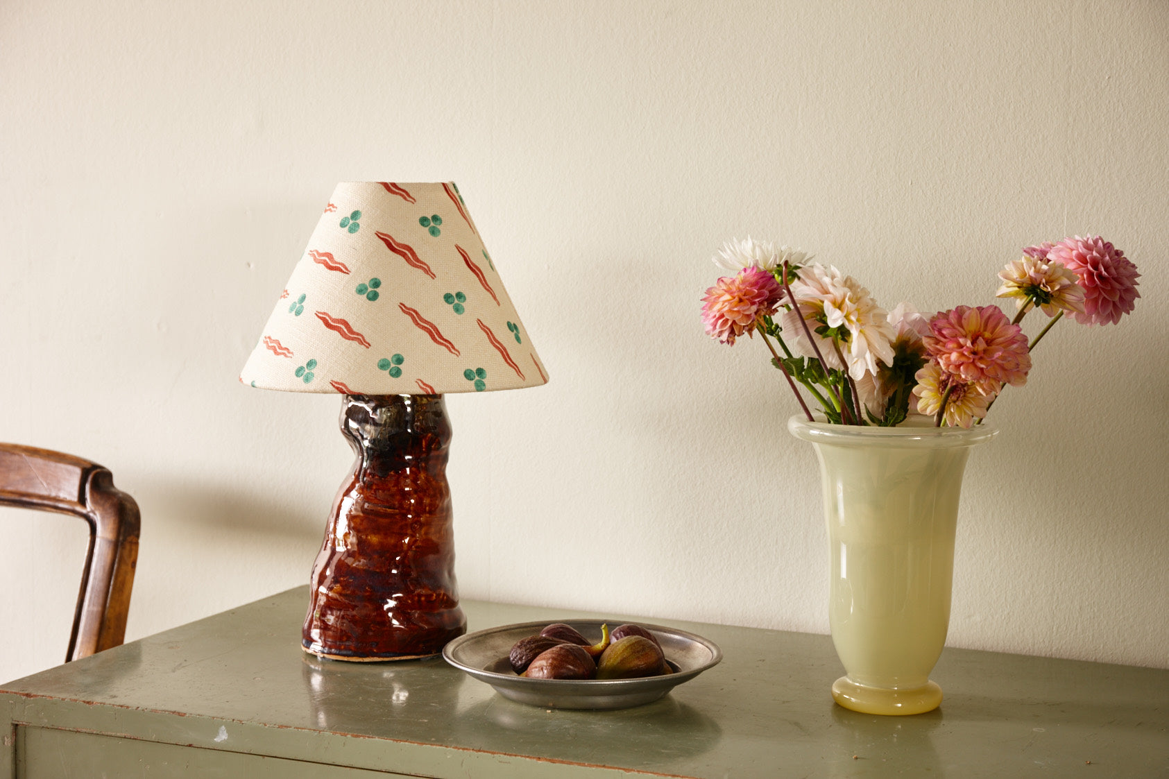A lamp with a patterned shade on a green cabinet, beside a bowl of figs and a vase of dahlias