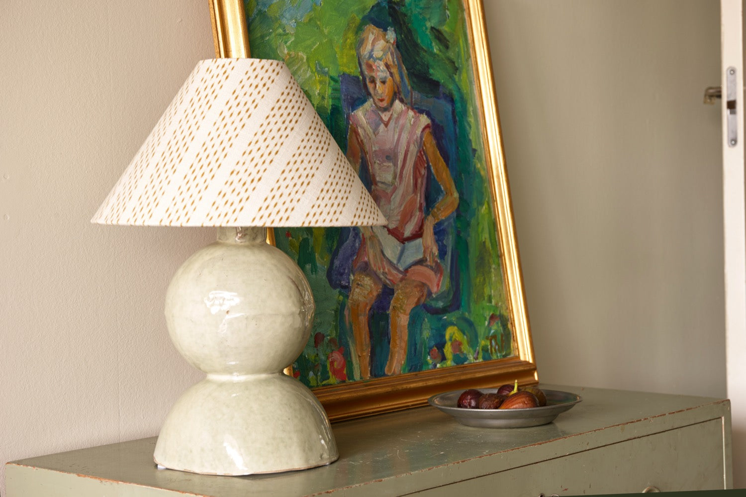A pale ceramic lamp with a diamond patterned shade on a green painted cabinet, beside a framed painting and a dish of figs
