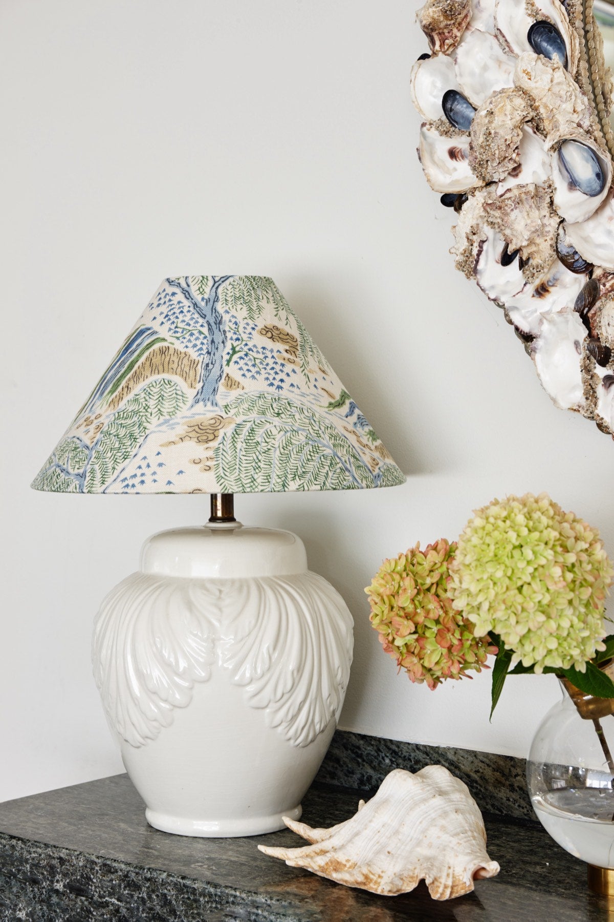 A lampshade printed with a willow landscape on a white ceramic base, on a stone counter beside hydrangeas and a conch shell