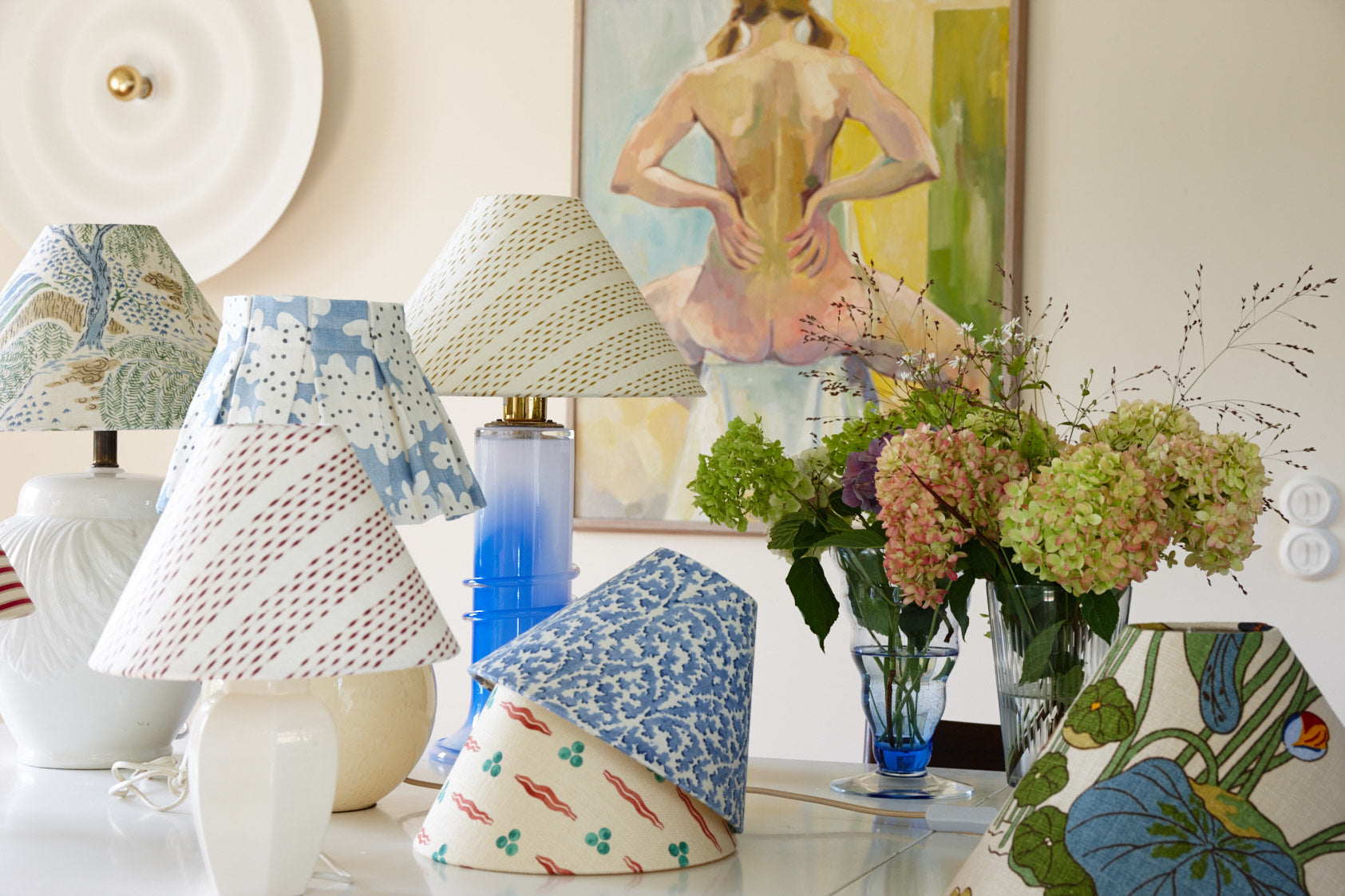 A group of lampshades in different patterns on a table, beside a vase of hydrangeas