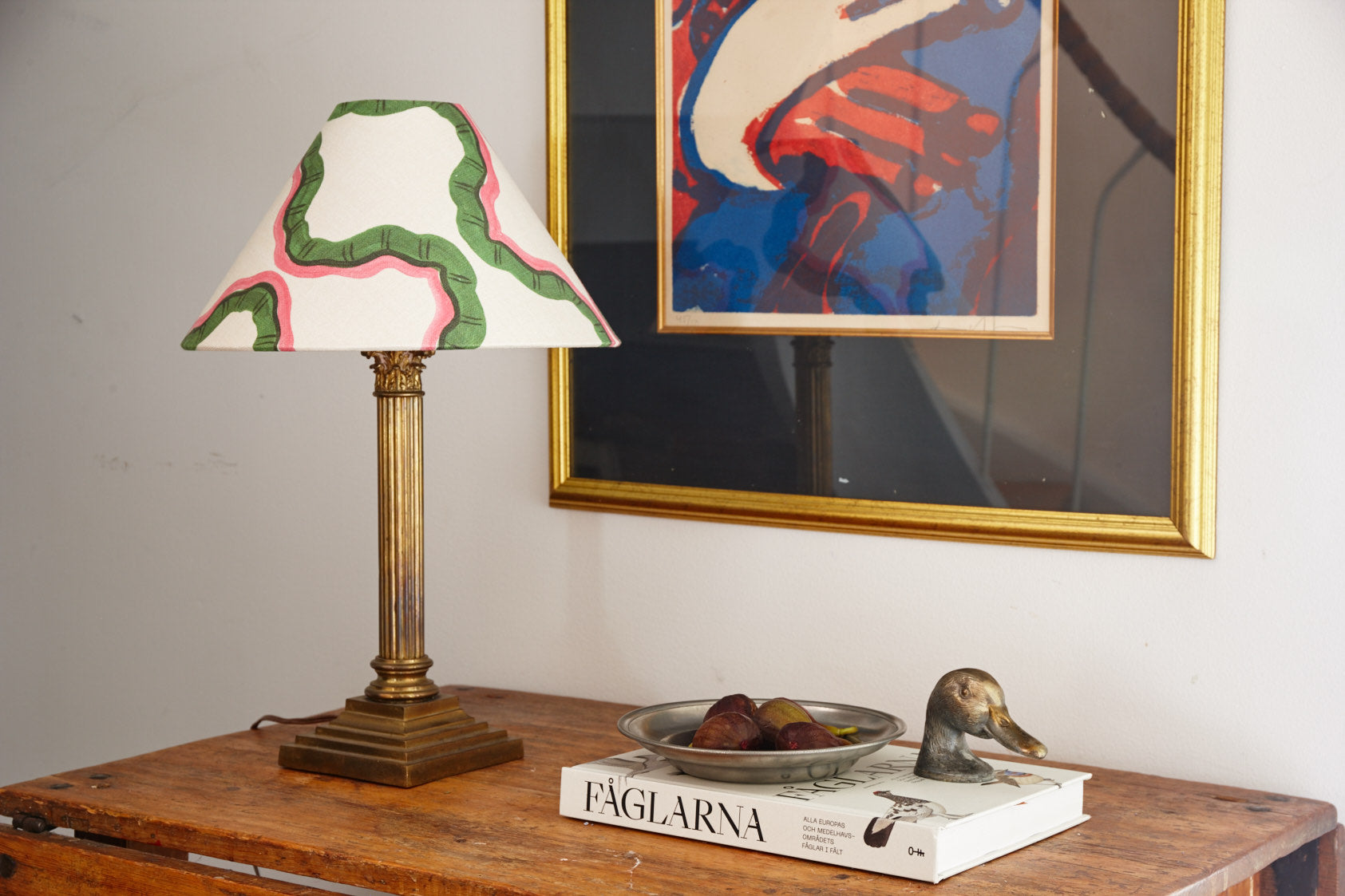 A table lamp with a hand painted green and pink shade on a wooden table, below a framed print