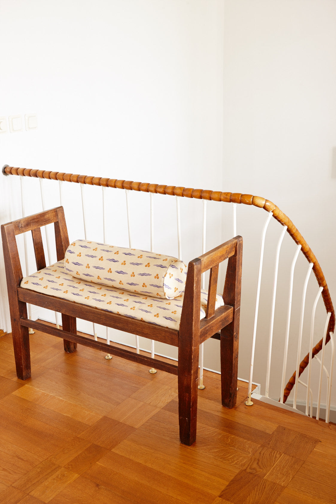 A wooden hall bench with a printed seat cushion and a bolster, beside a stair rail