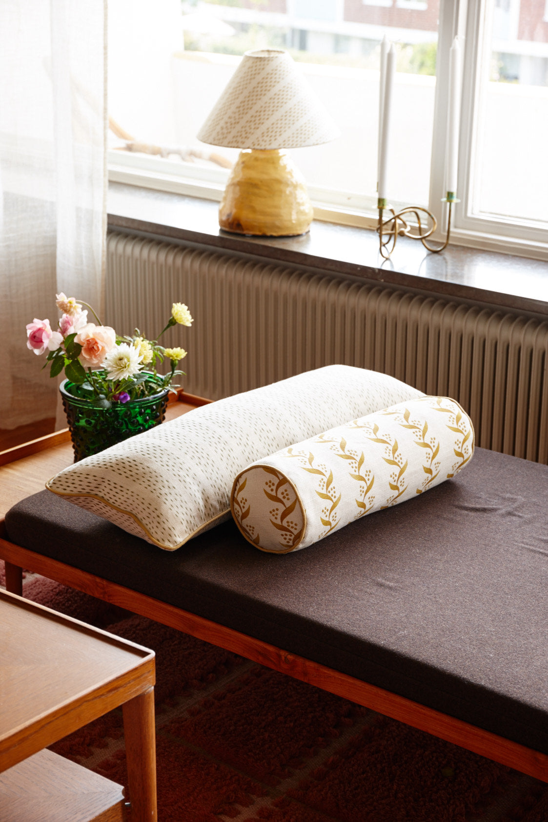A daybed with a long printed cushion and a bolster, beside a window with a lamp and flowers