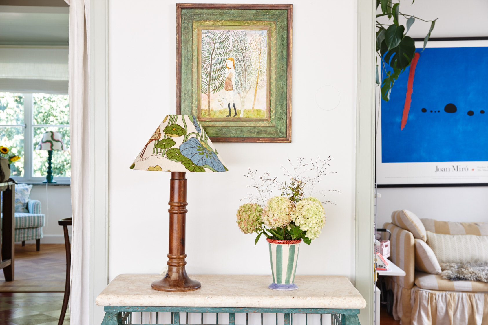 A lamp with a leaf patterned shade on a marble console table, beneath a framed painting, beside hydrangeas in a striped vase