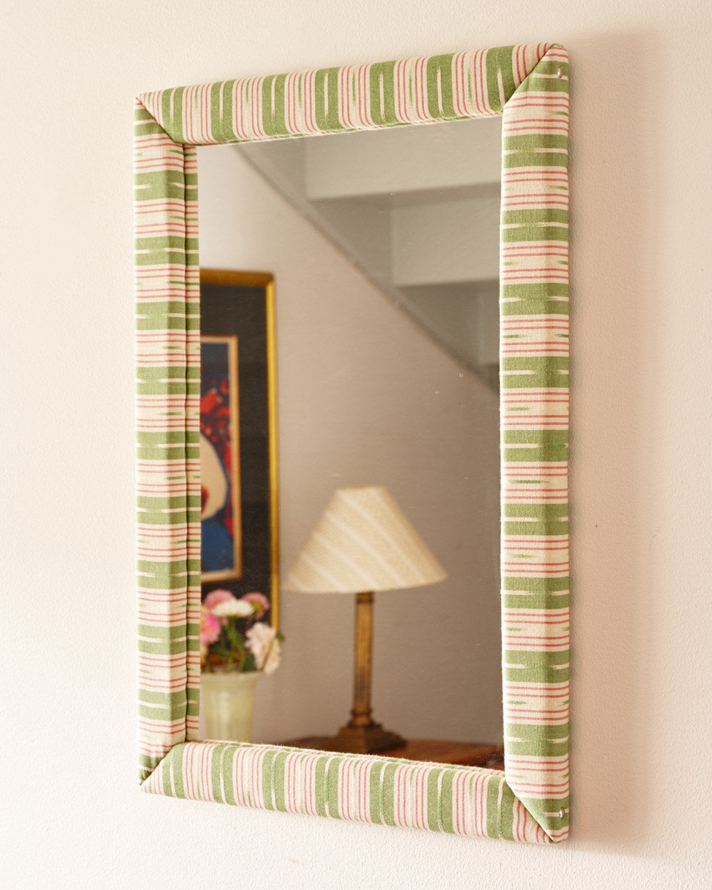 A mirror in a green and pink striped fabric on a wall, reflecting a lamp and a framed picture