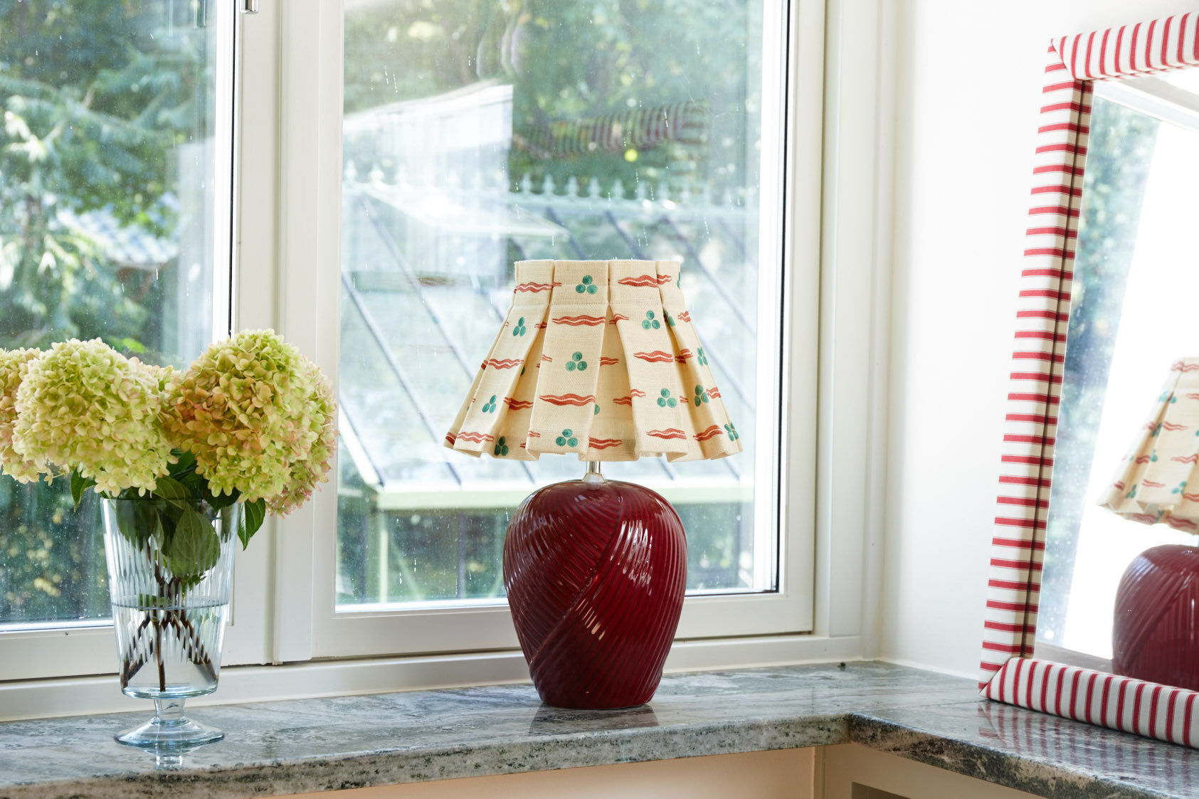 A mirror in red and white striped ticking beside a window, reflecting a table lamp with a box pleated shade