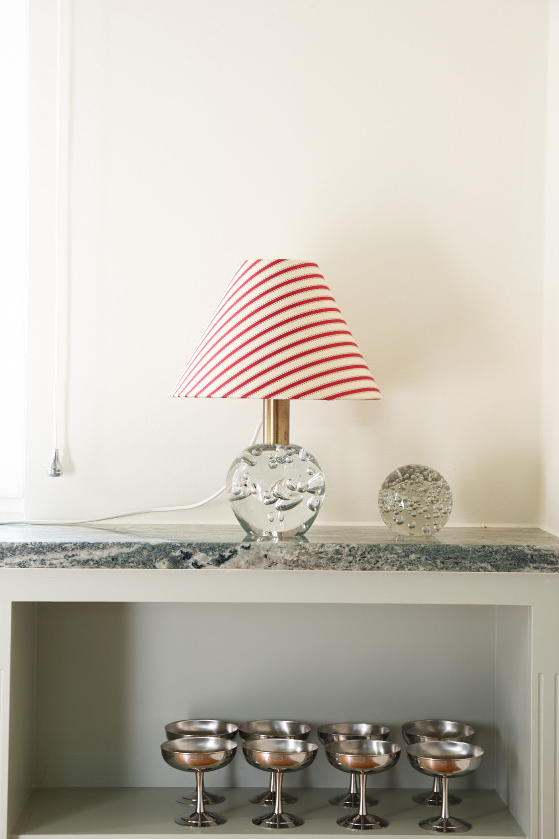 A lamp with a red and white striped shade on a bubbled glass base, on a marble shelf above a row of silver coupes