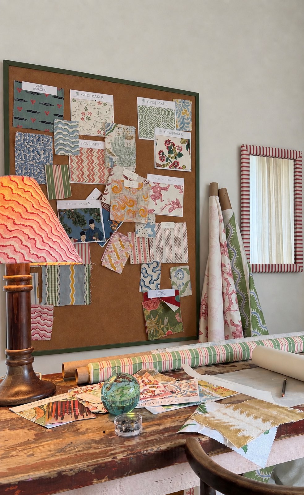 A pinboard of fabric swatches in a green frame, beside a striped mirror and rolls of printed fabric, above a work table of samples in the atelier