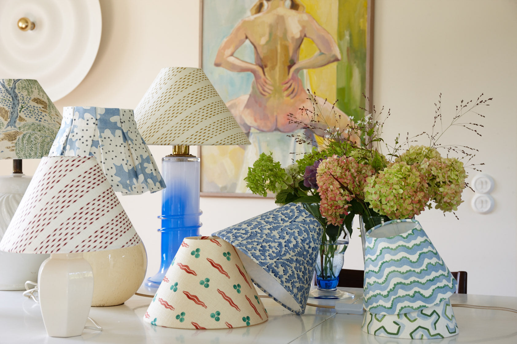 A group of lampshades in printed and painted cottons gathered on a table, with a vase of hydrangeas behind them