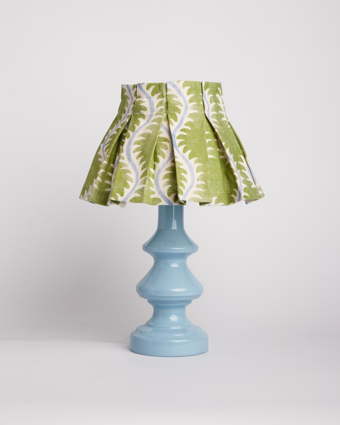 A box pleated lampshade in a green fern print on a turned pale blue base, against a plain studio backdrop