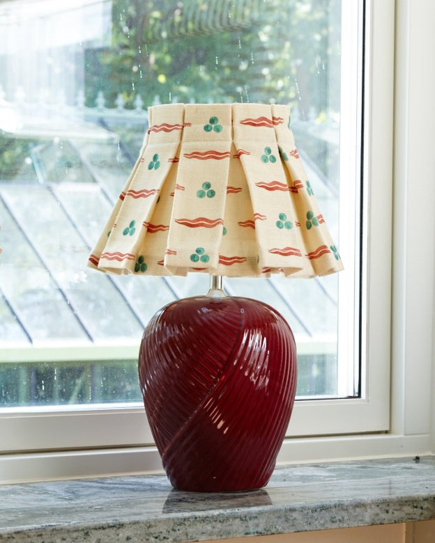 A box pleated lampshade in a cream print with red waves and green dots, on a ribbed deep red ceramic base in a window