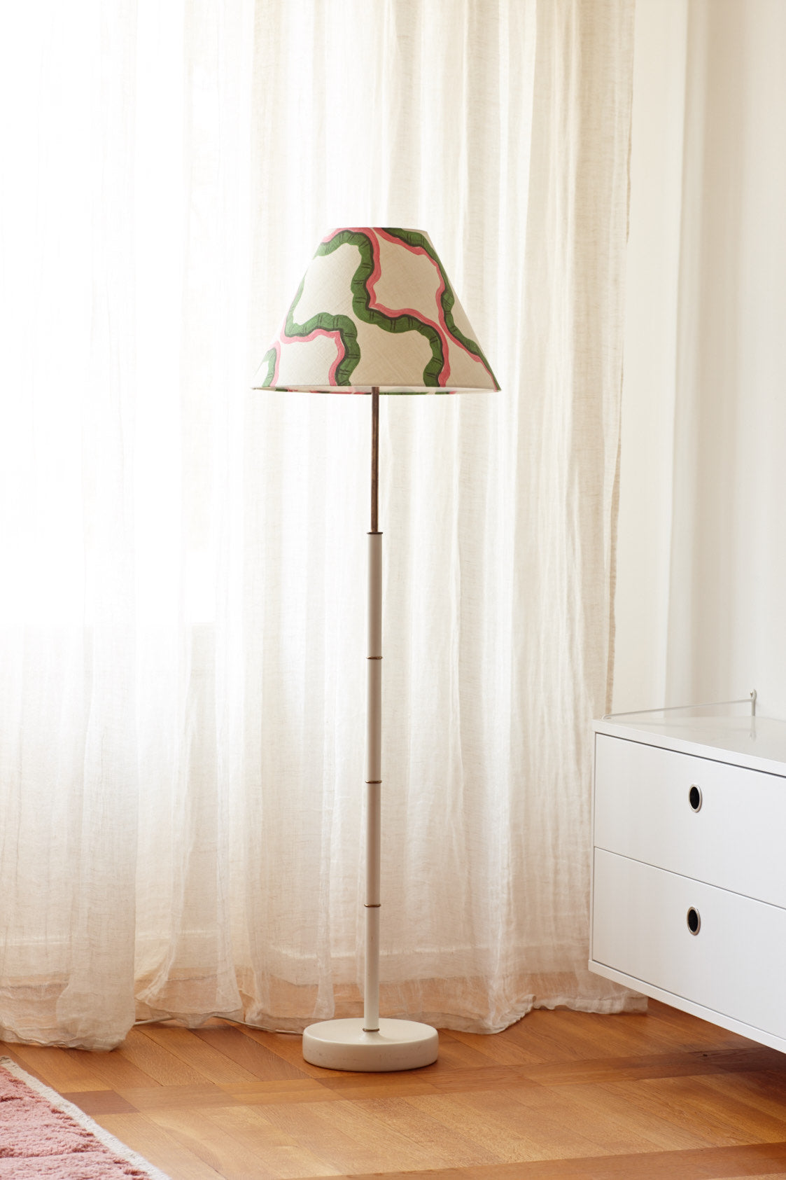 A floor lamp with a shade in a green and pink wave print, standing against sheer linen curtains beside a white chest of drawers