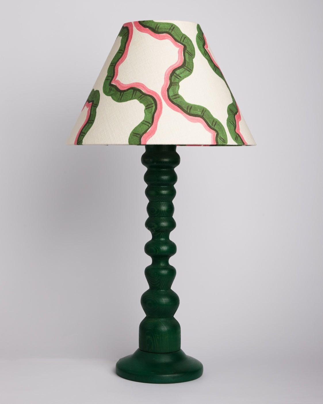 A lampshade in a green and pink wave print on a turned dark green wooden base, against a plain studio backdrop