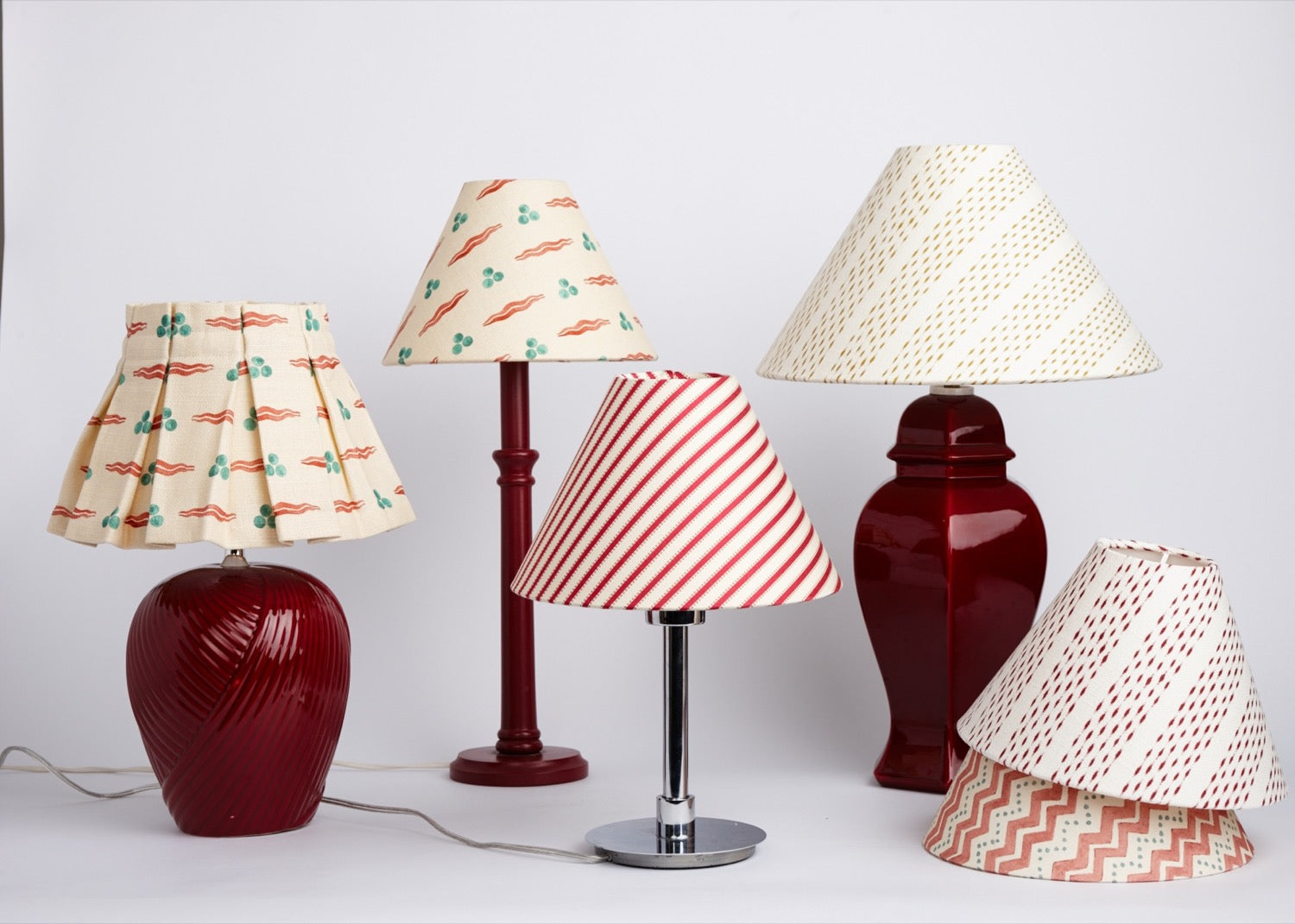 Five lampshades in different sizes and patterns grouped on a pale studio backdrop, two of them on deep red ceramic bases