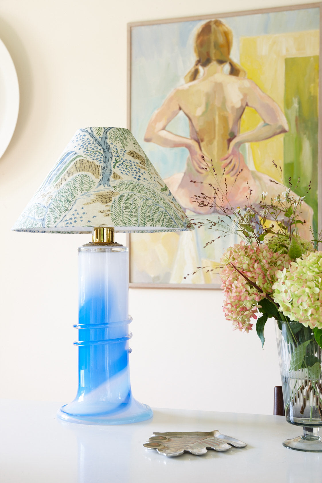 A lampshade in a green and blue landscape print on a swirled blue glass base, beside a vase of hydrangeas