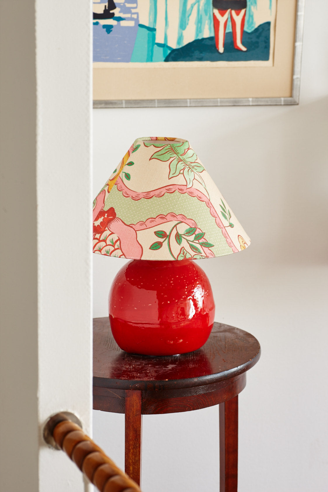 A lampshade in a pink and green scenic print on a round red ceramic base, on a dark wooden side table