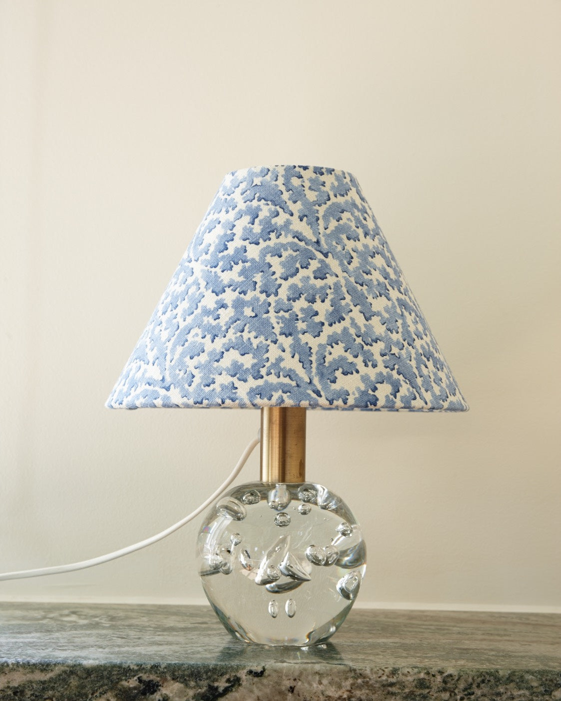 A lampshade in a blue leaf print on a clear bubbled glass base, standing on a green marble shelf