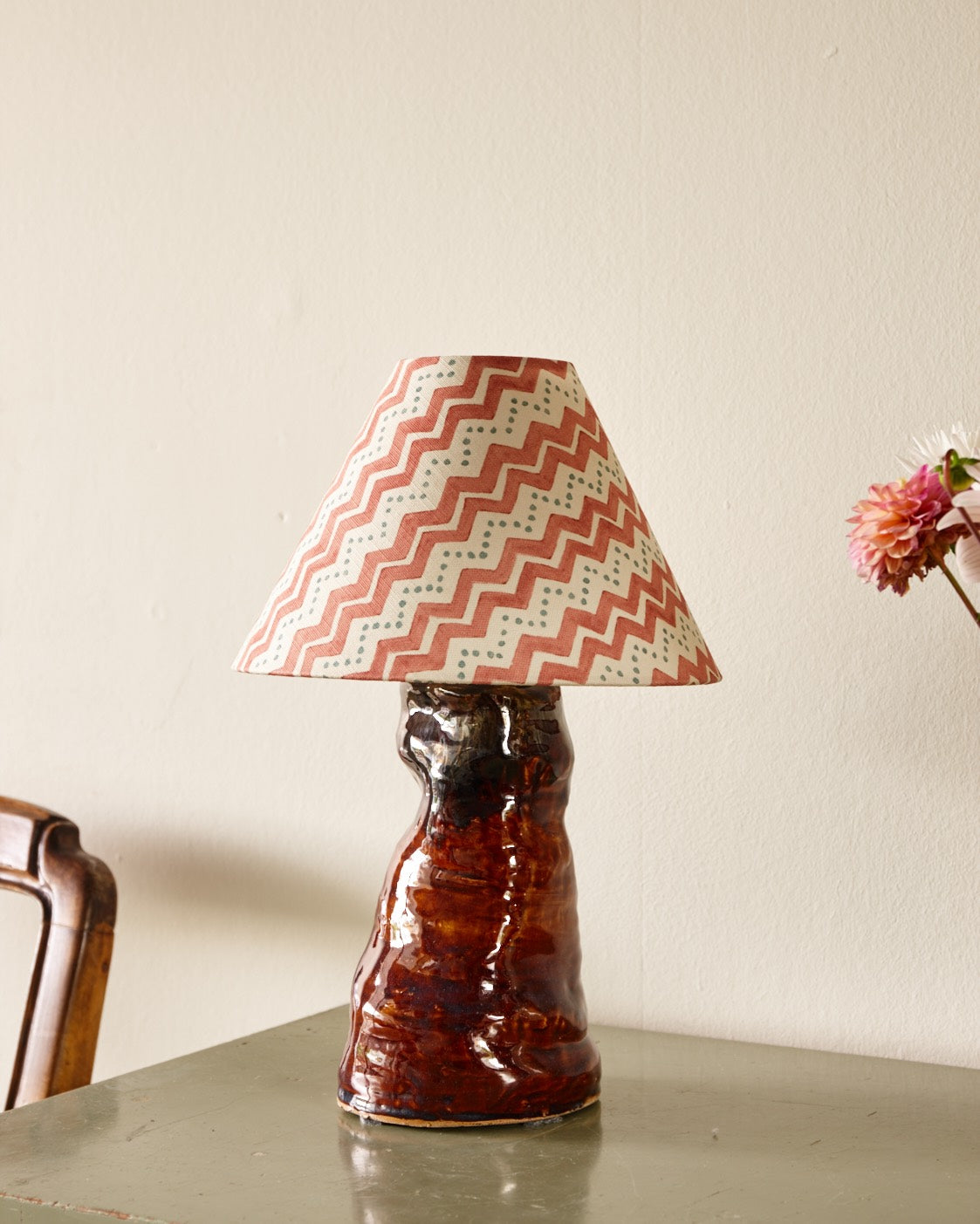 A lampshade in a red zigzag print on a glazed brown ceramic base, on a green painted table beside a dahlia