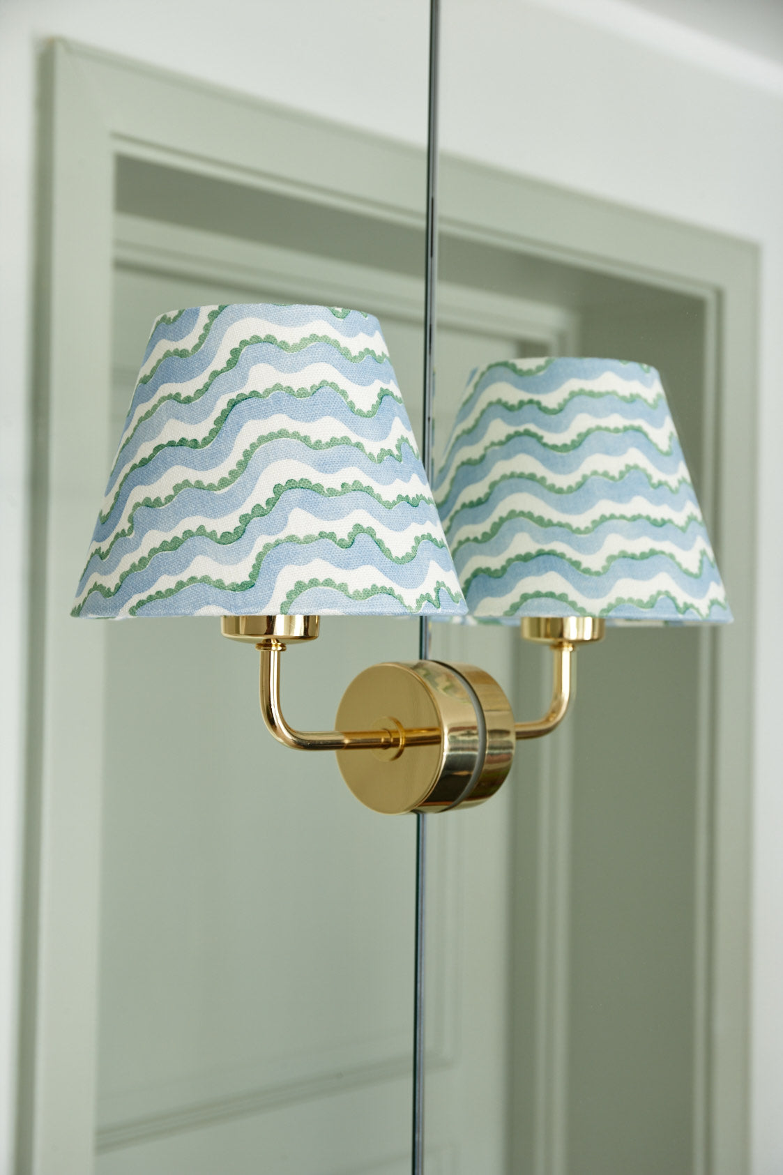 A brass wall light with two small shades in a blue and green wave print, reflected in a mirrored door