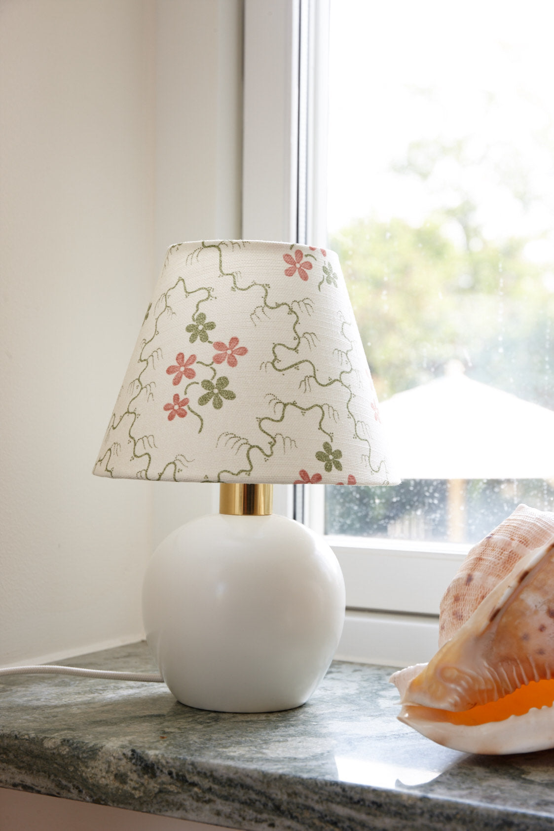 A small lampshade in a floral print on a round white base, standing on a marble windowsill beside a shell