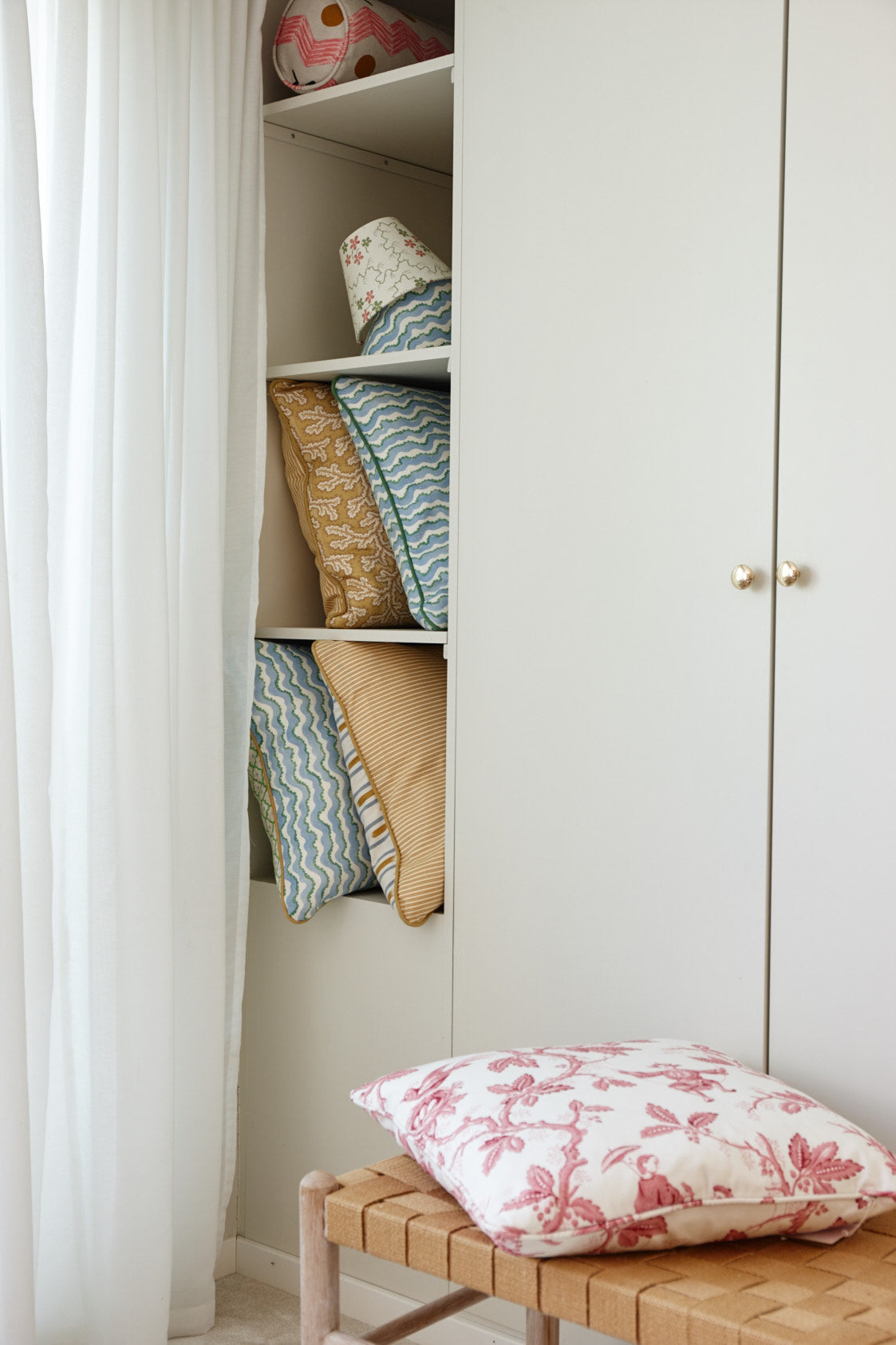 Cushions and a lampshade on the shelves of a cupboard, with a cushion on a woven bench